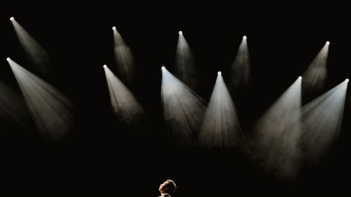 A lone performer on a dark stage beneath a bank of spotlights that are all aimed somewhere else