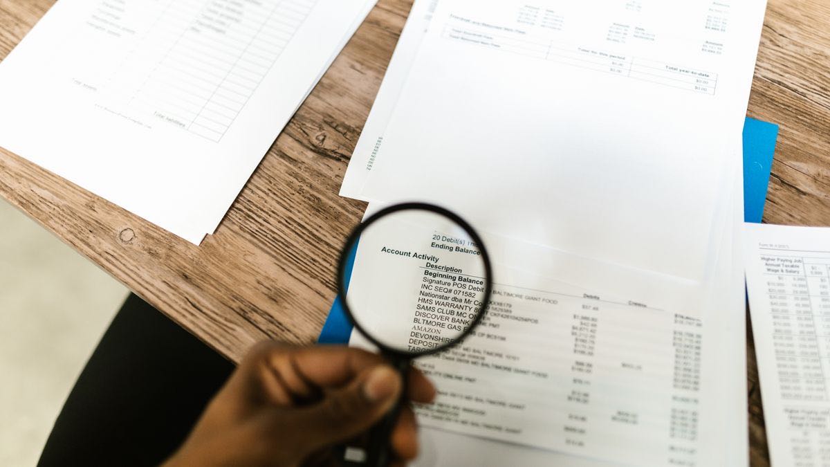 A hand holding a magnifying glass over financial statements on a desk, examining line items