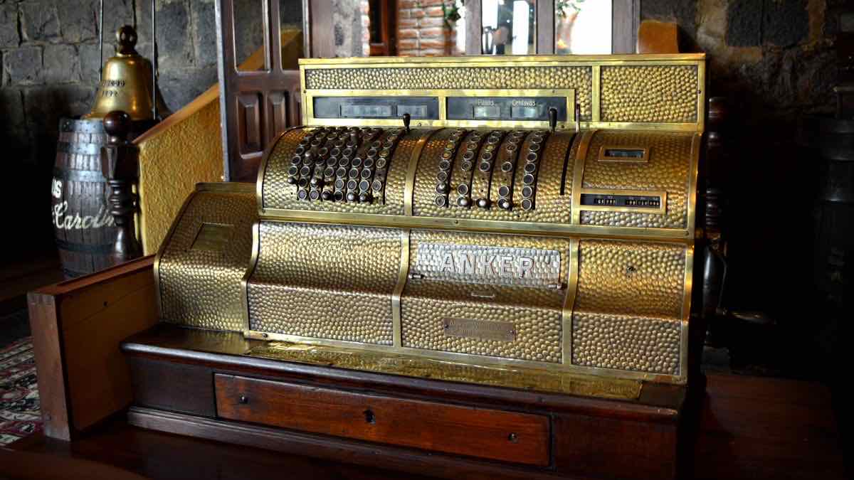 An ornate antique brass cash register on a wooden counter in a dim stone-walled room, with a brass bell and a wooden barrel behind it