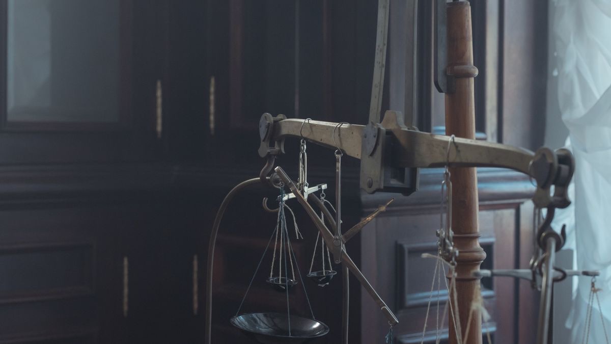 An antique two-pan brass balance scale hanging in a dark wood-panelled room, one pan lower than the other