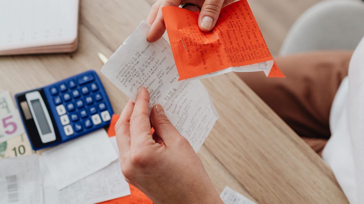 Two hands holding up paper till receipts over a wooden desk, with a blue pocket calculator and banknotes beside them