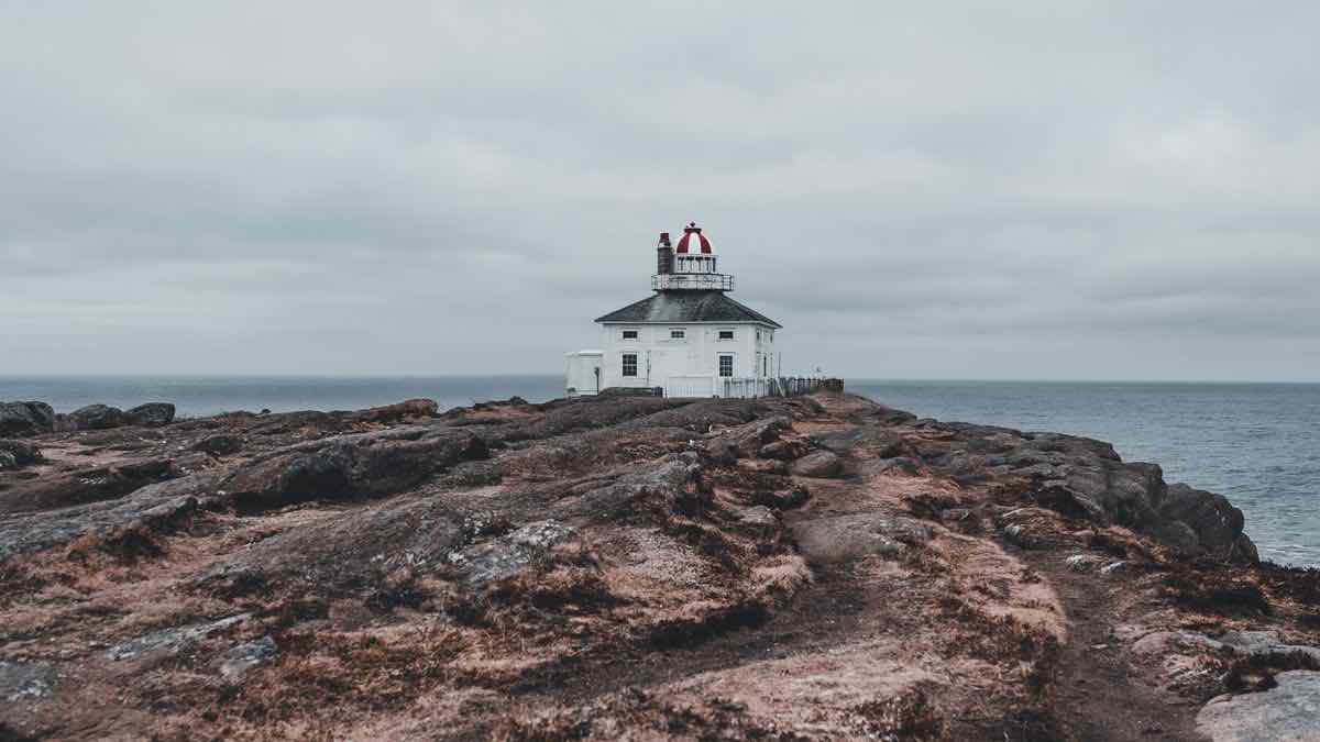 A white lighthouse keeper's house with a red lantern room, alone on a bare rocky headland under a flat grey sky
