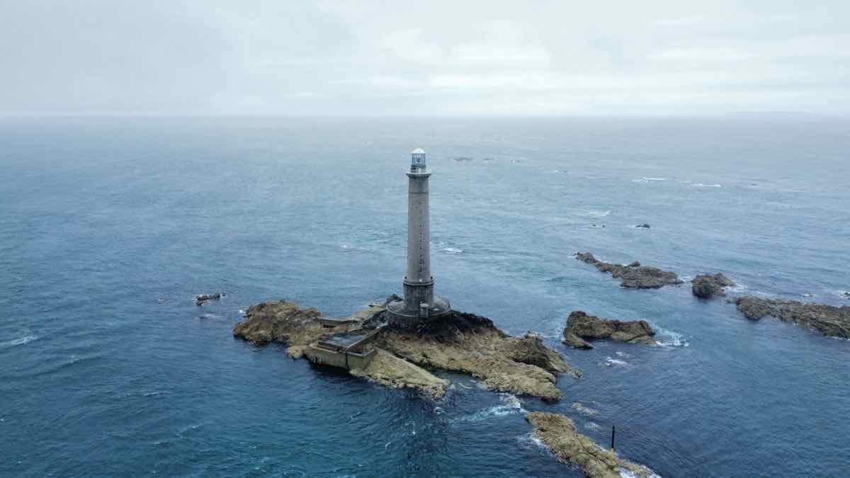 An aerial view of a single tall lighthouse on a small rocky outcrop, open grey sea all round it