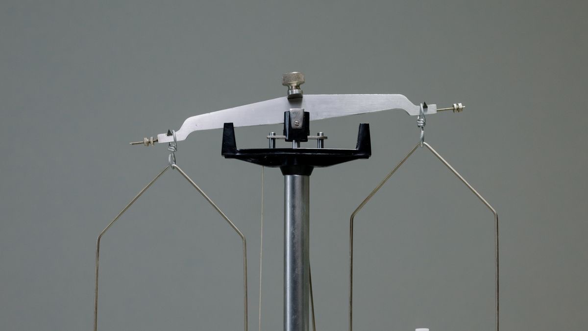 An antique balance scale photographed against a plain grey wall with both of its pans empty