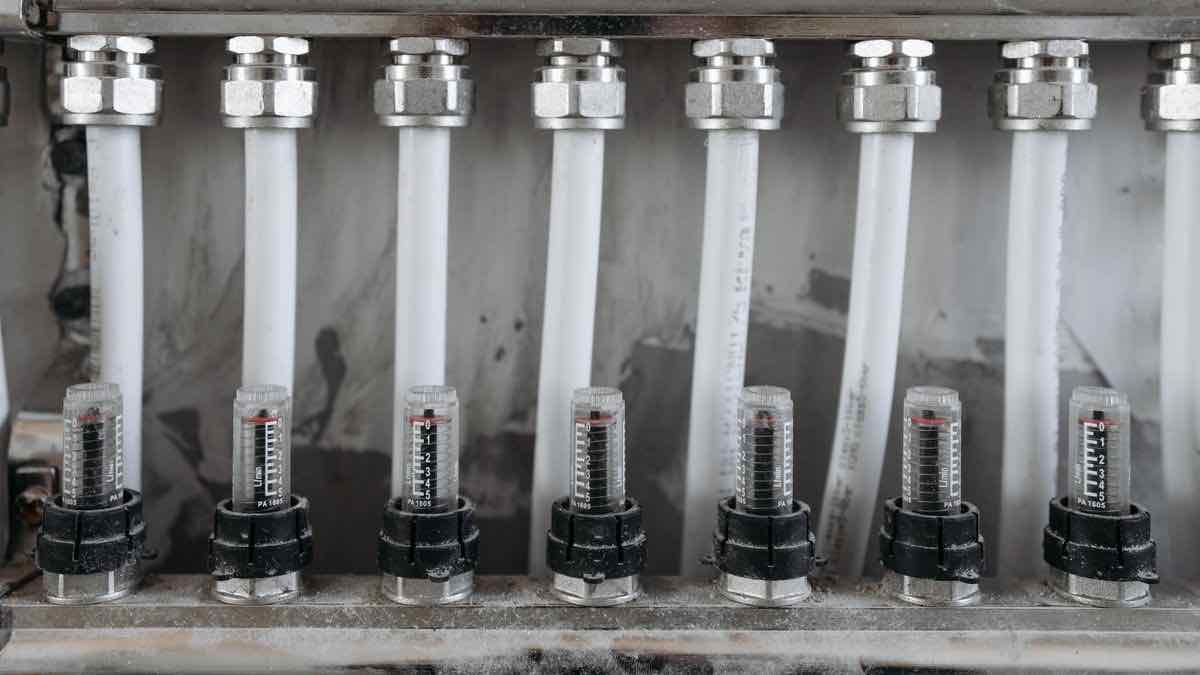 Eight identical white outlet pipes branching off one steel supply rail, each fitted with its own separate flow gauge
