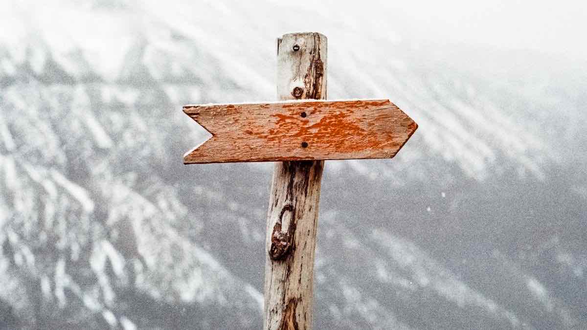 A weathered wooden arrow signpost pointing one direction against a snowy mountainside